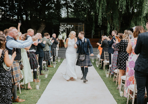 Bride and groom smile together as they walk back down the aisle at The Venue, Eskmills while their guests clap